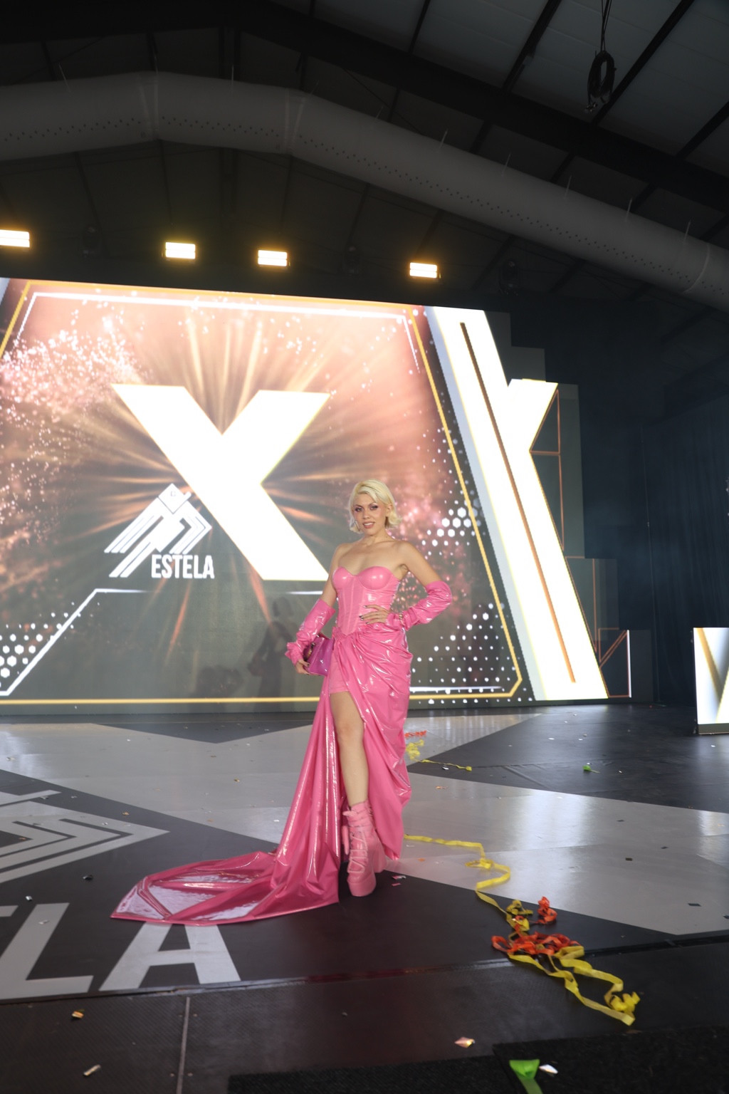 Suzette Labra de cuerpo completo con vestido corsé rosa, guantes y botas de plataforma, posando en un escenario ante un fondo iluminado de Estela X, con la cola del vestido visible